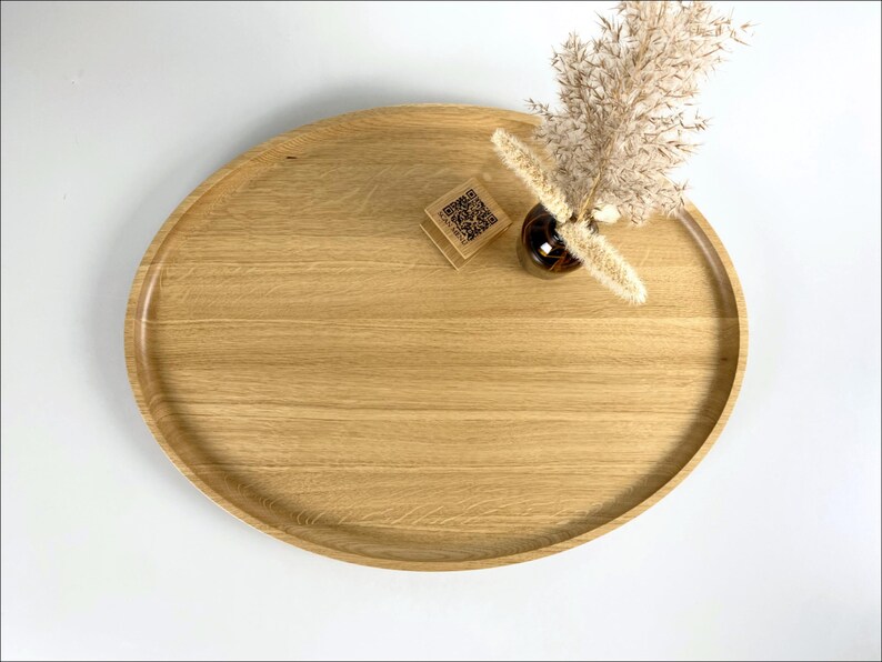 A minimalist wooden oval serving tray with a raised edge, featuring an amber glass bottle containing dried pampas grass and a small wooden block with a QR code placed on its surface. The tray is photographed against a plain white background.
