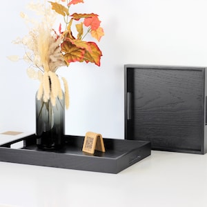 Two black wooden serving trays with handles, one displaying a smoky glass vase filled with autumn leaves and pampas grass, and a wooden QR menu stand. The second tray stands upright showing the textured wood grain finish. Shot on white background.