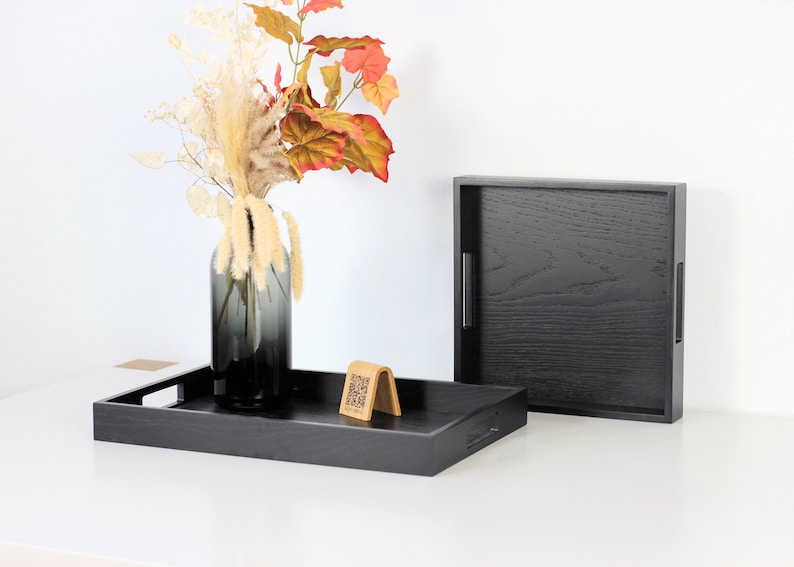 Two black wooden serving trays with handles, one displaying a smoky glass vase filled with autumn leaves and pampas grass, and a wooden QR menu stand. The second tray stands upright showing the textured wood grain finish. Shot on white background.