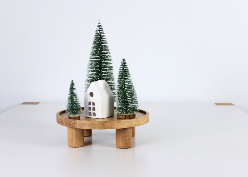 May include: A white ceramic house figurine sits on a wooden tiered stand with three small green artificial Christmas trees.