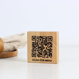 Op de afbeelding: Een houten blok met een QR-code en de tekst "SCAN FOR MENU" gegraveerd. Het blok is gemaakt van lichtgekleurd hout en is vierkant. Een houten servetring en een opgerold servet staan op de achtergrond.