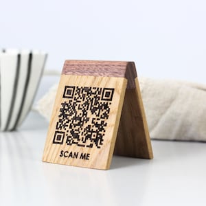 Puede incluir: Un letrero de código QR de madera con base marrón claro y parte superior marrón oscuro. El letrero está grabado con un código QR y las palabras "SCAN ME". El letrero está de pie.