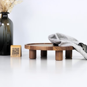 以下が含まれることがあります： 丸い天板と4本の短い脚が付いた木製のサービングトレイ。灰色の布がトレイに掛けられています。「SCAN MENU」というテキストとQRコードが付いた木製のブロックが左側にあります。背景には、ドライフラワーが入った濃い色のガラスの花瓶があります。