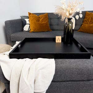 May include: A black wooden serving tray with a rectangular shape and handles. The tray is placed on a grey ottoman, with a cream blanket draped over it. A black vase with dried flowers and a wooden block with the letter "A" are also visible.