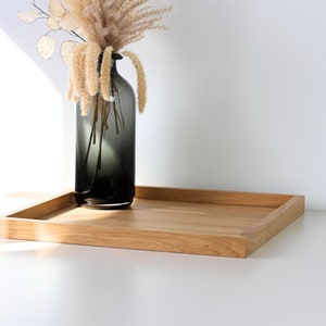 Könnte beinhalten: Ein rechteckiges Holztablett mit hellbrauner Maserung und erhöhtem Rand. Eine hohe, dunkelgraue Glasvase mit Farbverlauf steht auf dem Tablett und enthält ein getrocknetes Blumenarrangement mit beigen Federn und runden Stielen.