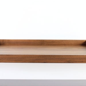 Puede incluir: Una bandeja rectangular de madera con un acabado de madera natural. La bandeja tiene un borde elevado y dos peque&ntilde;as asas recortadas en los lados.