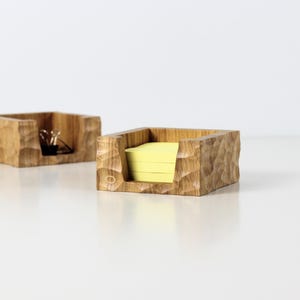 May include: Two wooden desk organizers with a textured, carved design. One contains yellow sticky notes, the other has paper clips and a binder clip. The organizers are square-shaped and made of light brown wood.