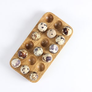 Pode incluir: Um suporte de ovos de madeira com 18 ranhuras, cheio de ovos de codorniz. Os ovos são salpicados com manchas marrons e brancas.