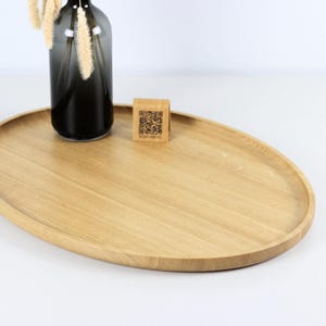 A minimalist wooden oval serving tray with a raised edge, featuring an amber glass bottle containing dried pampas grass and a small wooden block with a QR code placed on its surface. The tray is photographed against a plain white background.