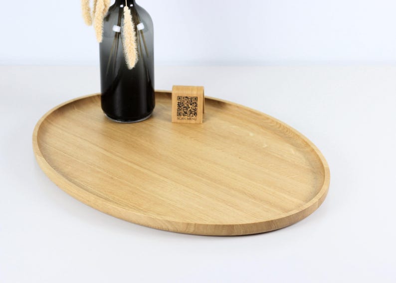 A minimalist wooden oval serving tray with a raised edge, featuring an amber glass bottle containing dried pampas grass and a small wooden block with a QR code placed on its surface. The tray is photographed against a plain white background.