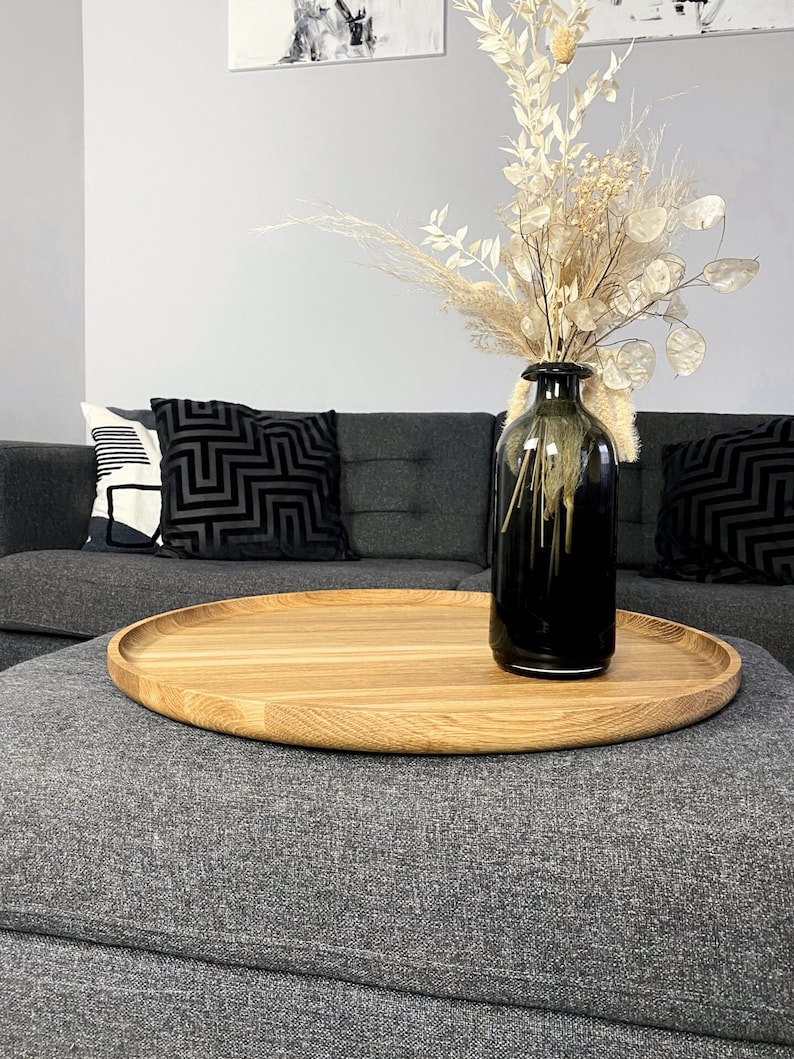Round oak wooden tray styled on a grey ottoman with a black vase and dried floral arrangement, in a modern living room.