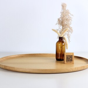 A minimalist wooden oval serving tray with a raised edge, featuring an amber glass bottle containing dried pampas grass and a small wooden block with a QR code placed on its surface. The tray is photographed against a plain white background.