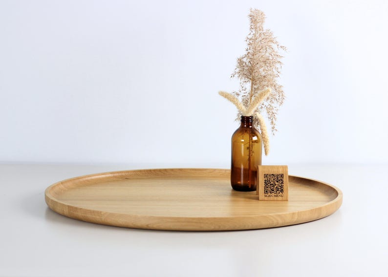 A minimalist wooden oval serving tray with a raised edge, featuring an amber glass bottle containing dried pampas grass and a small wooden block with a QR code placed on its surface. The tray is photographed against a plain white background.