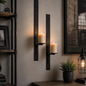 May include: Two black metal wall sconces with lit cream-colored candles. The sconces are mounted on a gray wall, with a dark wooden table and framed artwork in the background. A small potted plant and a caged light fixture are also visible.