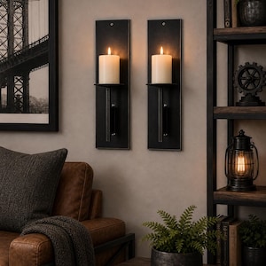 May include: Two black metal wall sconces with lit ivory candles. The sconces are mounted on a neutral wall, with a framed black and white photograph of a bridge to the left. A leather sofa and wooden shelves are also visible.