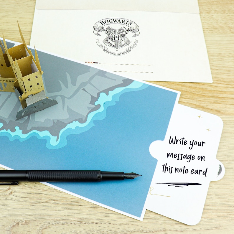 May include: A pop-up greeting card featuring a tan paper castle structure, a blue and gray illustrated landscape, and a white note card with the black text "Write your message on this note card." A black fountain pen and a Hogwarts envelope are included.