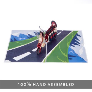 Puede incluir: Una scooter roja y blanca con una rueda negra, conduciendo por un camino con una zona verde y monta&ntilde;as en el fondo. La scooter es una tarjeta pop-up 3D.