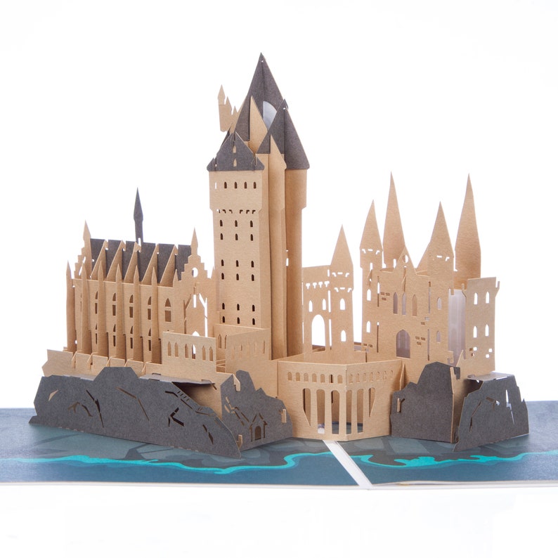 May include: A three-dimensional pop-up card featuring a detailed paper craft model of a castle. The structure is constructed from tan and dark gray cardstock, showcasing multiple towers, pointed spires, and arched windows, set upon a dark blue base.