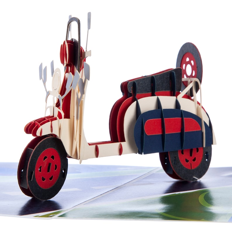 Puede incluir: Un modelo de papel de una scooter vintage roja, blanca y azul, con un portaequipajes y una rueda de repuesto. La scooter est&aacute; hecha de papel y tiene un efecto 3D.