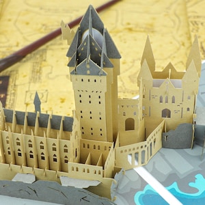 May include: A three-dimensional paper craft model of a castle featuring tan walls, grey roofs, and intricate architectural details including arched windows and tall towers. The structure is constructed from layered cardstock with cutouts.