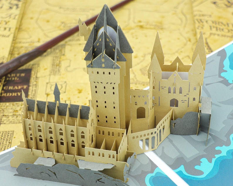 May include: A three-dimensional paper craft model of a castle featuring tan walls, grey roofs, and intricate architectural details including arched windows and tall towers. The structure is constructed from layered cardstock with cutouts.
