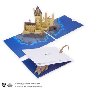 May include: A pop-up greeting card featuring a three-dimensional paper model of a castle on a rocky island surrounded by blue water. The card is shown both open and partially closed, with a brown arrow pointing to the white cover of the card.