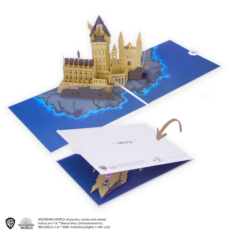 May include: A pop-up greeting card featuring a three-dimensional paper model of a castle on a rocky island surrounded by blue water. The card is shown both open and partially closed, with a brown arrow pointing to the white cover of the card.