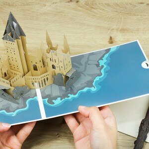 May include: A pop-up book featuring a tan paper castle structure with dark gray roofs, set against a blue and teal illustrated water base. Beside the open book lies a dark brown wand and a cream-colored card with the Hogwarts crest and text.