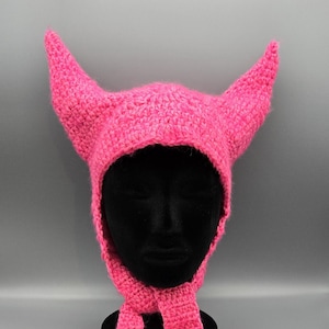 Puede incluir: Un gorro de ganchillo rosa brillante con dos cuernos puntiagudos y largas cintas colgantes. El gorro se exhibe sobre una cabeza de maniquí negra sobre un fondo gris. Un accesorio hecho a mano único.