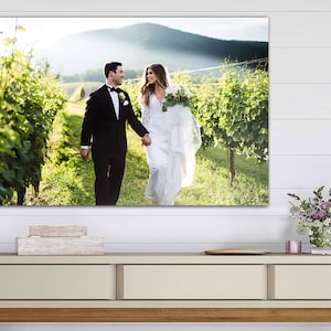 K&ouml;nnte beinhalten: Ein Hochzeitspaar geht Hand in Hand durch einen Weinberg, die Braut in einem wei&szlig;en Kleid und der Br&auml;utigam in einem schwarzen Anzug. Das Paar l&auml;chelt und schaut sich an.