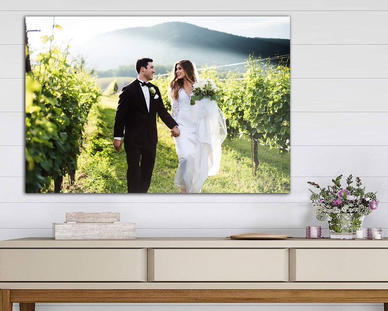 K&ouml;nnte beinhalten: Ein Hochzeitspaar geht Hand in Hand durch einen Weinberg, die Braut in einem wei&szlig;en Kleid und der Br&auml;utigam in einem schwarzen Anzug. Das Paar l&auml;chelt und schaut sich an.