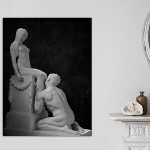 May include: A black and white photograph of two nude sculptures. The female sculpture is standing on a pedestal and the male sculpture is kneeling at her feet.