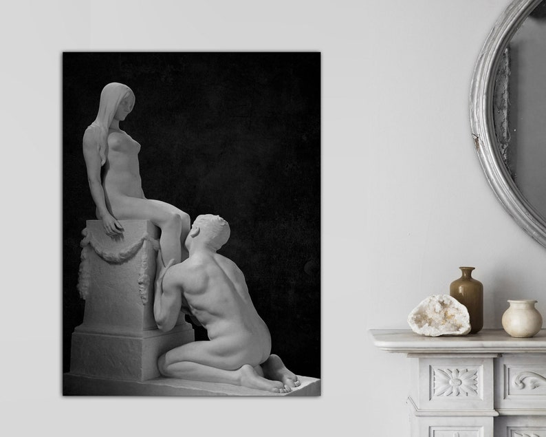 May include: A black and white photograph of two nude sculptures. The female sculpture is standing on a pedestal and the male sculpture is kneeling at her feet.
