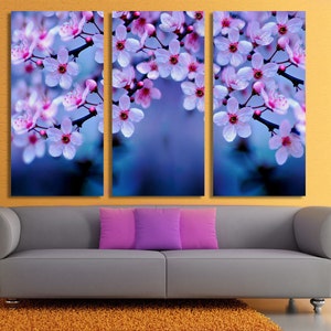May include: A triptych canvas art piece featuring close-up images of cherry blossoms. The blossoms are white with pink centers and are set against a blurred blue background. The artwork is displayed above a gray sofa with purple pillows.