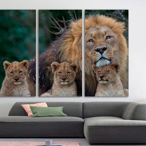 May include: A large male lion with a thick mane is surrounded by three young cubs. The lion is looking directly at the camera, while the cubs are looking at the lion. The background is a blurred green forest.