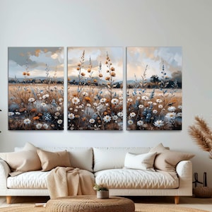 Op de afbeelding: Een drieluik canvas kunstprint met een sereen veld vol wilde bloemen. Het kunstwerk toont witte, gele en blauwe bloemen tegen een achtergrond van een zachte, bewolkte lucht. Het kleurenpalet is gedempt met warme tinten.