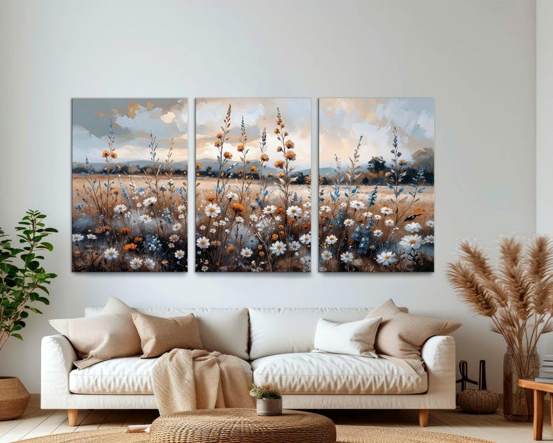 Op de afbeelding: Een drieluik canvas kunstprint met een sereen veld vol wilde bloemen. Het kunstwerk toont witte, gele en blauwe bloemen tegen een achtergrond van een zachte, bewolkte lucht. Het kleurenpalet is gedempt met warme tinten.
