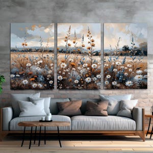 Op de afbeelding: Een drieluik van canvas kunst met een aquarel schilderij van een veld met wilde bloemen. Het kunstwerk toont witte, gele en blauwe bloemen tegen een achtergrond van een veld en een bewolkte lucht. De kunst wordt boven een grijze bank getoond.