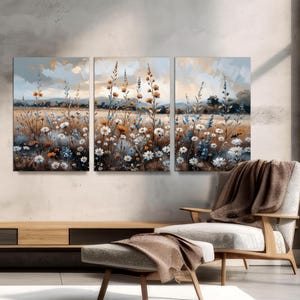 Op de afbeelding: Een driedelig canvas kunstwerk dat een veld met wilde bloemen in aquarel stijl toont. Het kunstwerk toont witte, blauwe en oranje bloemen met een zacht, gedempt kleurenpalet. De achtergrond toont een landschap met een bewolkte lucht.