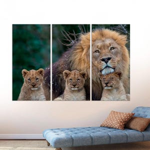 May include: A large male lion with a thick mane is surrounded by three cubs. The lion is looking directly at the camera, while the cubs are looking at the lion. The background is a blurred green forest.