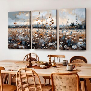 Op de afbeelding: Een drieluik van canvas kunst met een veld vol wilde bloemen in tinten wit, oranje en blauw. Het kunstwerk heeft een zachte, impressionistische stijl met een gedempte kleurenpalet. De schilderijen worden boven een houten eettafel tentoongesteld.