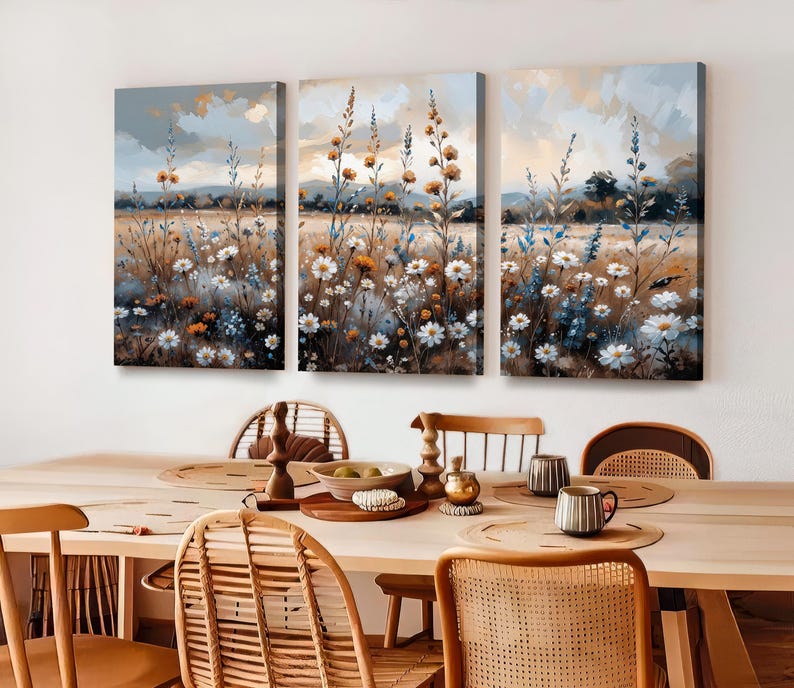 Op de afbeelding: Een drieluik van canvas kunst met een veld vol wilde bloemen in tinten wit, oranje en blauw. Het kunstwerk heeft een zachte, impressionistische stijl met een gedempte kleurenpalet. De schilderijen worden boven een houten eettafel tentoongesteld.
