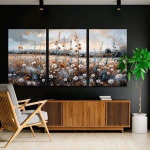 Op de afbeelding: Een driedelig canvas kunstwerk met een veld met wilde bloemen in wit, oranje en blauwe tinten. Het schilderij heeft een zachte aquarel stijl met een gedempte kleurenpalet. De achtergrond toont een bewolkte lucht en verre heuvels.