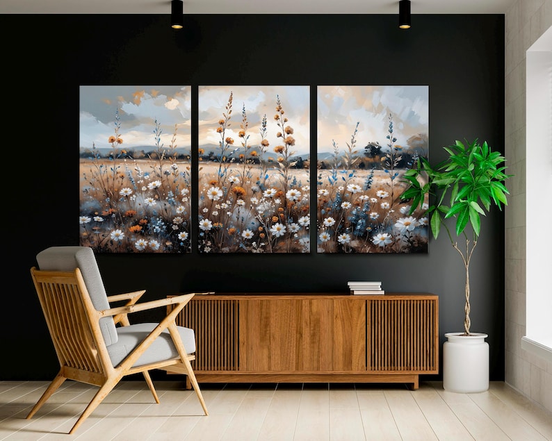 Op de afbeelding: Een driedelig canvas kunstwerk met een veld met wilde bloemen in wit, oranje en blauwe tinten. Het schilderij heeft een zachte aquarel stijl met een gedempte kleurenpalet. De achtergrond toont een bewolkte lucht en verre heuvels.