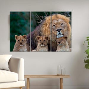 May include: A large male lion with a thick mane stands with three cubs. The lion is looking directly at the camera. The cubs are nestled against the lion's body. The background is a blurred green forest.