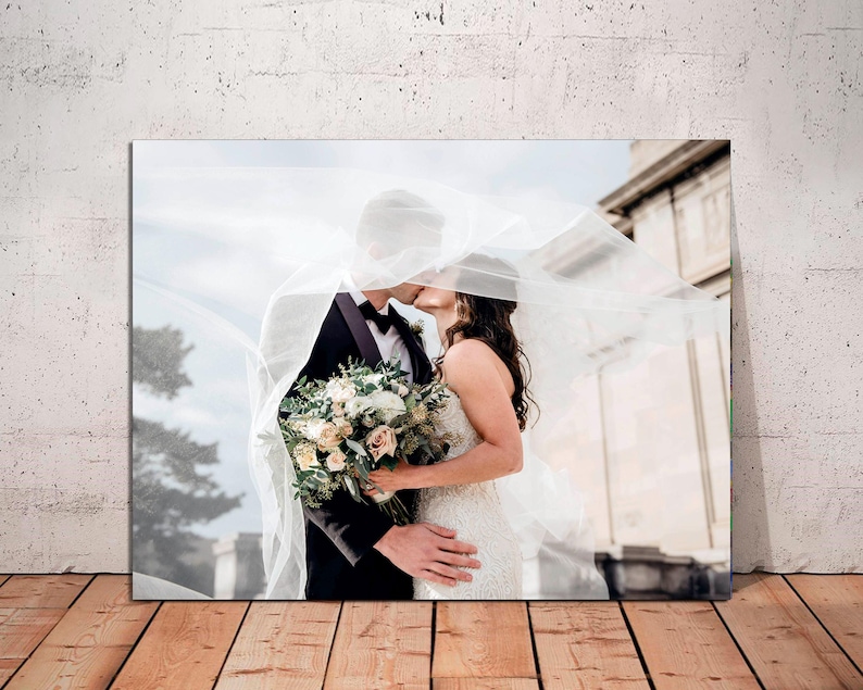 May include: A bride and groom kiss under a white veil, with a bouquet of white and green flowers in the bride's hand.