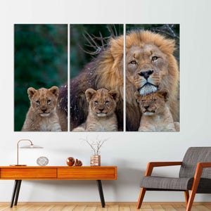 May include: A large male lion with a thick mane stands with two young cubs nestled against him. The lion is brown and has a light brown mane. The cubs are light brown with dark brown spots. The background is a blurred green forest.