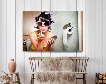 May include: A woman with curlers and sunglasses applies lipstick in a bathroom setting. She wears a yellow top and holds a small mirror. A roll of toilet paper hangs on the wall behind her.
