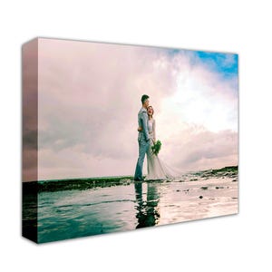 May include: A couple pose for a wedding photograph on a beach. The bride is wearing a white dress and the groom is wearing a blue suit. The sky is cloudy and the water is calm.