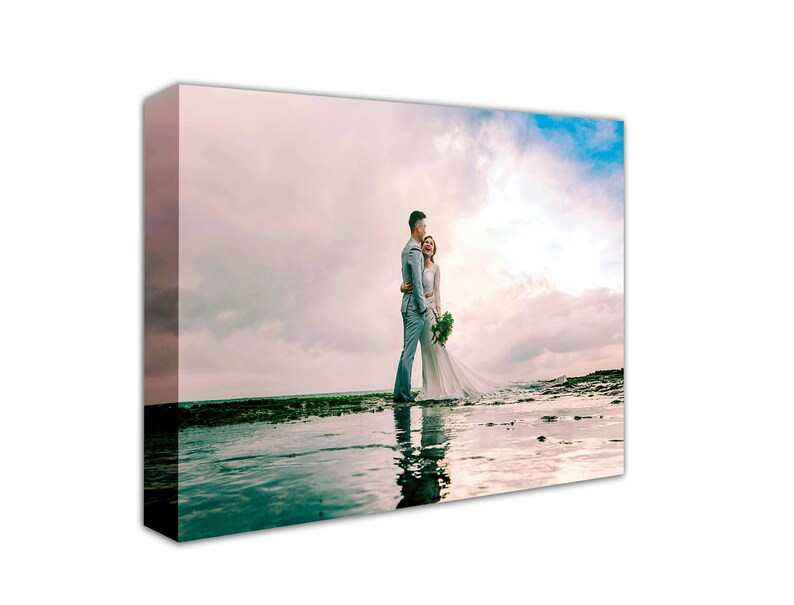 May include: A couple pose for a wedding photograph on a beach. The bride is wearing a white dress and the groom is wearing a blue suit. The sky is cloudy and the water is calm.